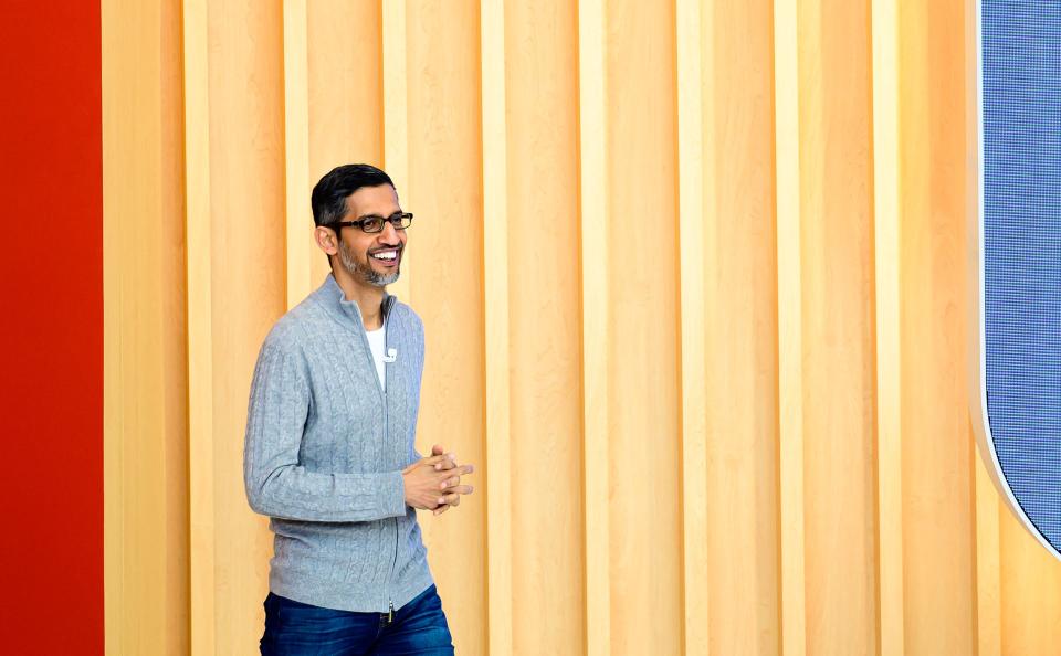 Sundar Pichai walking onto a stage with a yellow-striped background while wearing a grey long-sleeve top.