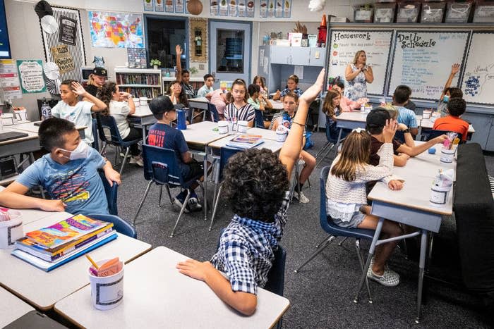 A classroom full of kids with some holding their hands up and some wearing masks