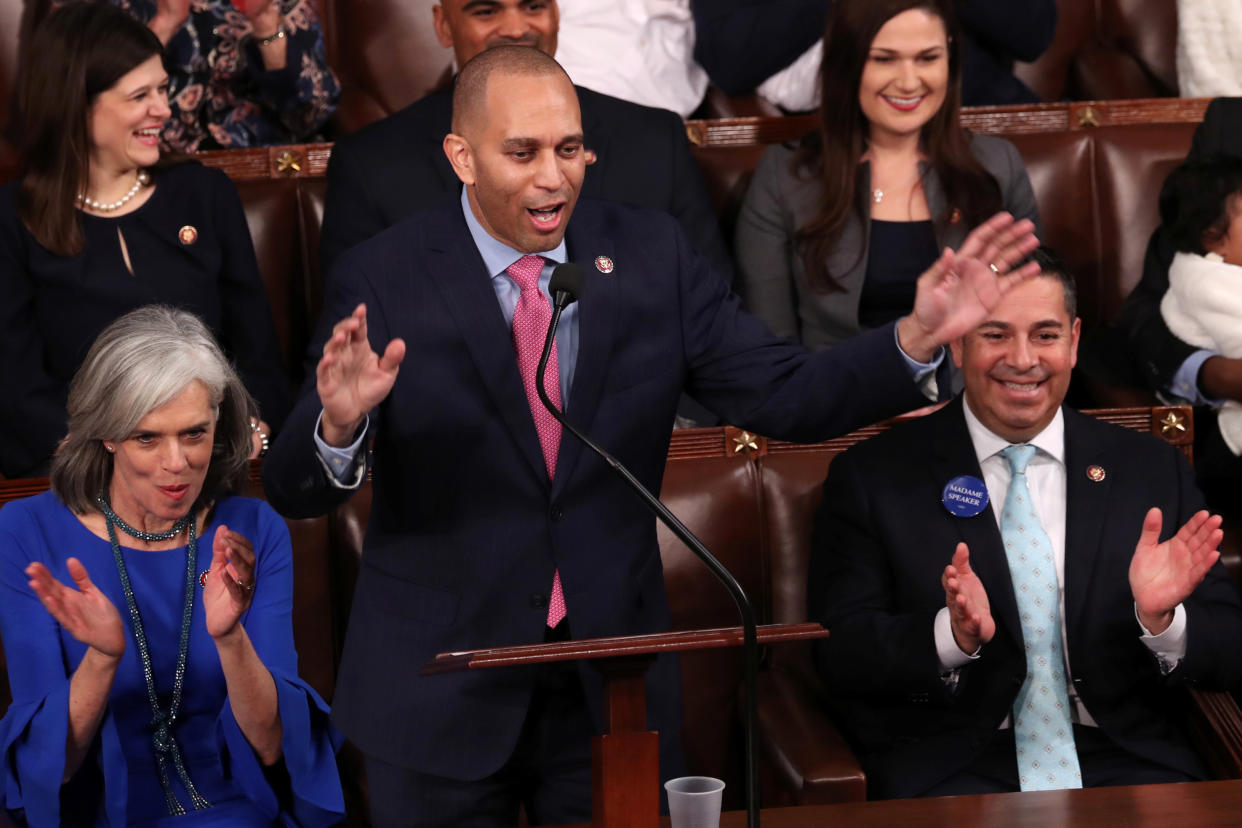 Hakeem Jeffries stands at a podium with a microphone with his hands held up, surrounded by a half dozen others who are seated and apparently applauding.