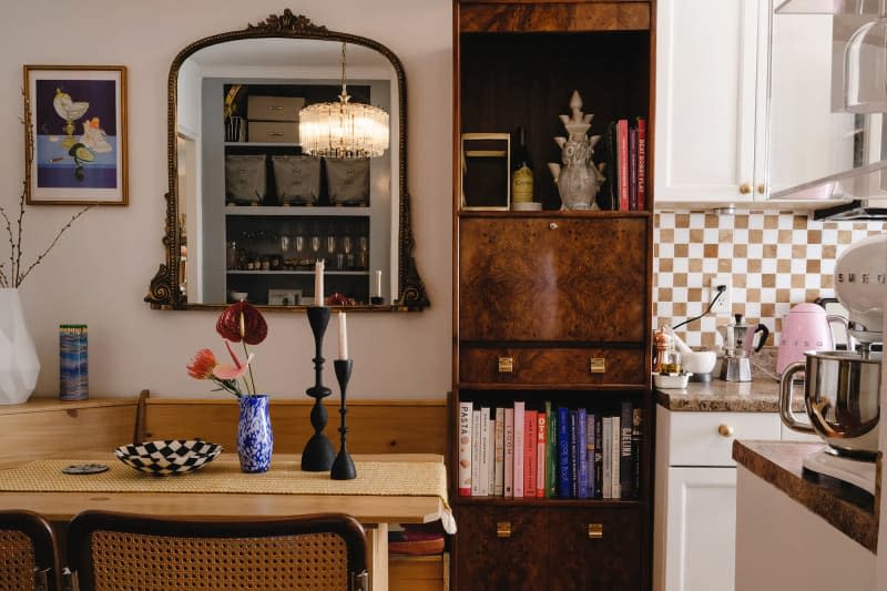 Dining room with light wood table, rattan chairs with checkered tan and white cushions, bronze ornate framed mirror, tall wood cabinet/bookshelf, modern curved white shelf on wall with art objets, fringed chandelier, vase with flowers, candles with black candlesticks on table