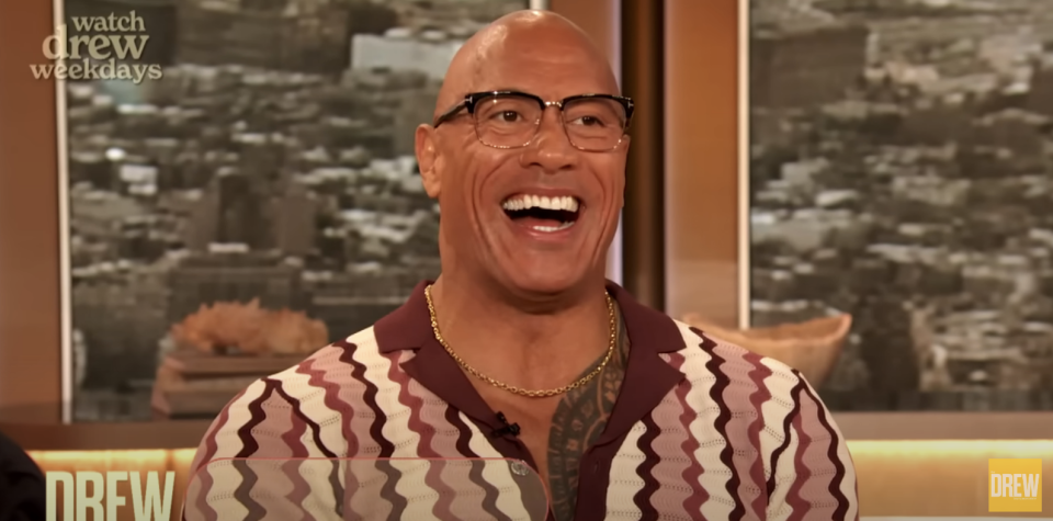 Dwayne Johnson laughing during a talk show interview, wearing a striped shirt and a necklace