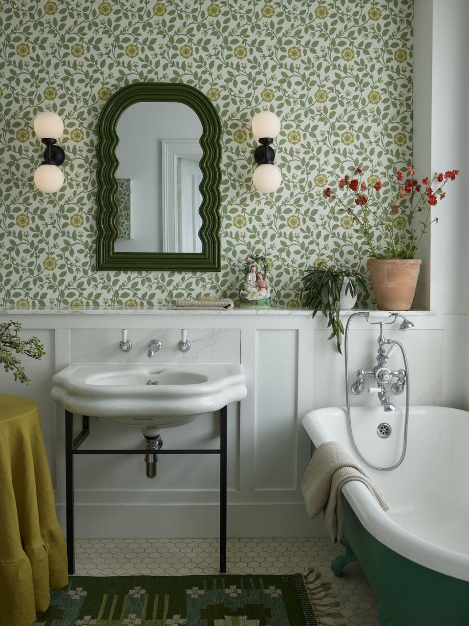 a wallpapered bathroom with panelling, shelving, a bath, sink and green mirror