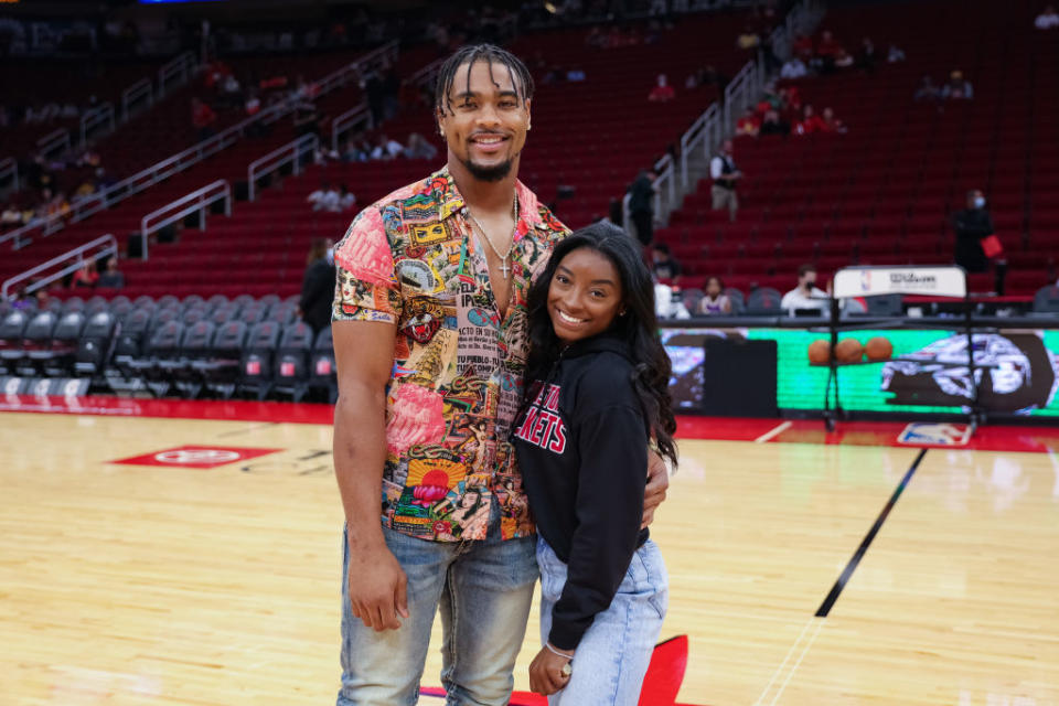Simone Biles and Jonathan Owens are standing on a basketball court, smiling at the camera. Jonathan is wearing a patterned shirt and jeans, Simone is in a sweatshirt and jeans