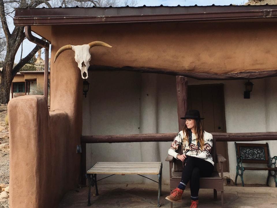 Emily Hart sits in a chair, while wearing a black fedora, a white patterned sweater, black pants, and boots. She is sitting in an outdoor area with a bull skull on the wall.