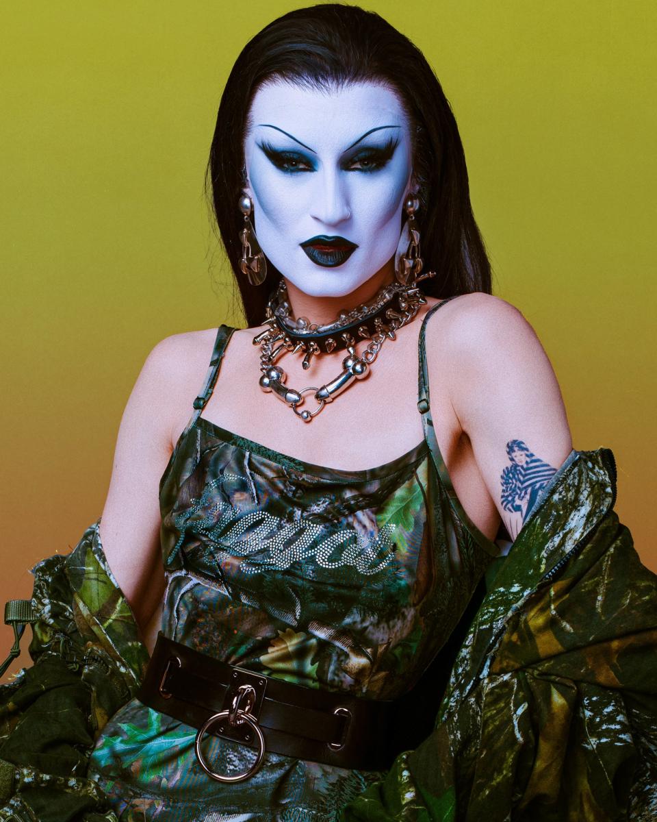 Gottmik in a green patterned dress and heavy makeup, featuring dark lipstick, white face makeup, and dramatic eye makeup, stands against a yellow background
