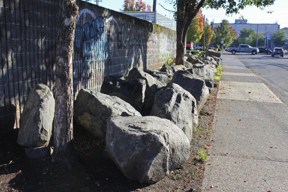 Roughly 30 large boulders occupy the narrow strip of land between a sidewalk and a parking lot wall in Portland, Ore., on Oct. 17, 2023. The boulders were installed sometime after late July at the site of a former homeless encampment to prevent tents from being set back up. The encampment was cleared several times over the course of the year. Cities across the U.S. are struggling with and cracking down on tent encampments as the number of homeless people grows, largely due to a lack of affordable housing. Homeless people and their advocates say sweeps are cruel and costly, and there aren't enough homes or beds for everyone. (AP Photo/Claire Rush)