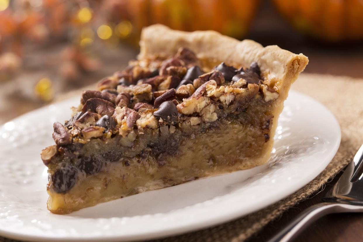 A slice of chocolate pecan pie on a plate with a burlap place setting and fork.  In the background is Thanksgiving / Fall decorations.  The decorations are a variety of pumpkins and gourds.  The shot is horizontal with selective focus.