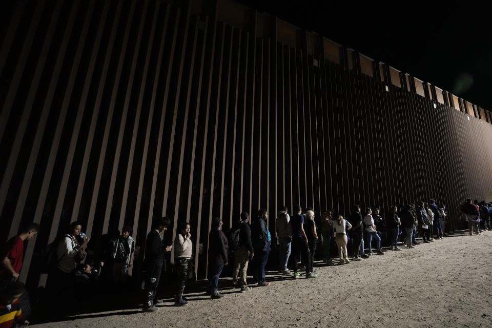 Un grupo de personas hacen fila junto al muro fronterizo mientras esperan para solicitar asilo tras cruzar al frontera desde México, el 11 de julio de 2023, cerca de Yuma, Arizona. El repunte en la inmigración ha sido posible por el descubrimiento este año de una nueva a través de Nicaragua, donde la laxitud de los requisitos de entrada permite a los mauritanos y a otros extranjeros adquirir una visa de bajo costo sin necesidad de demostrar que van a continuar su viaje. (AP Foto/Gregory Bull)