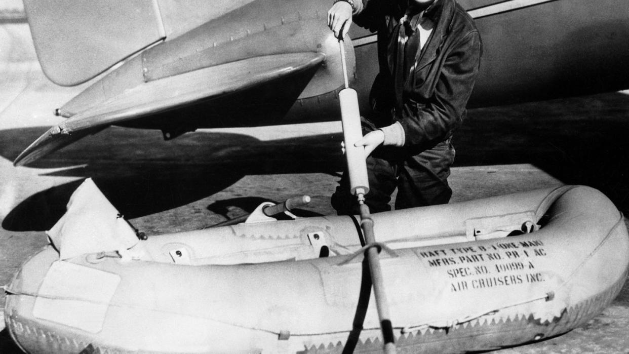 amelia earhart smiles while holding an oar and standing between an inflatable lifeboat and a plane, she wears a dark jumpsuit