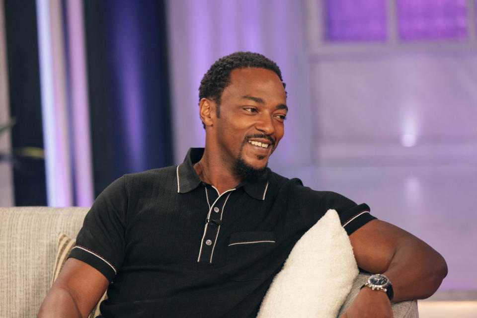 Anthony Mackie smiling while seated on a couch, wearing a casual black polo shirt