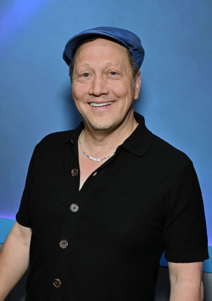 Rob Schneider lächelt und trägt ein schwarzes Hemd mit Knöpfen und eine blaue Kappe vor blauem Hintergrund