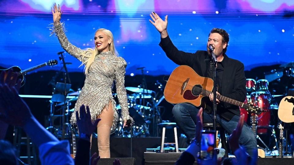 Gwen Stefani in a sparkly silver dress waves to the crowd in sync with husband Blake Shelton sitting on a stool with his guitar at the Power of Love Gala