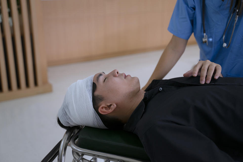 A person with a bandaged head lies on a stretcher, attended by a healthcare professional wearing a blue uniform and a stethoscope