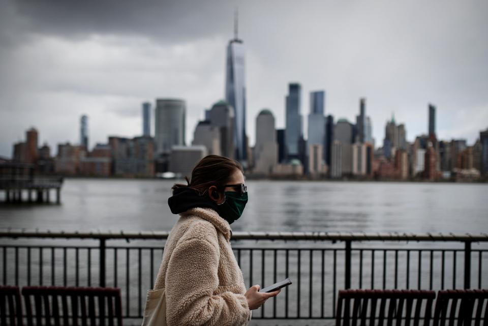 In this April 10, 2020, file photo a woman wearing a face mask sure to COVID-19 concerns walks along the Jersey City waterfront with the New York City skyline in the background.