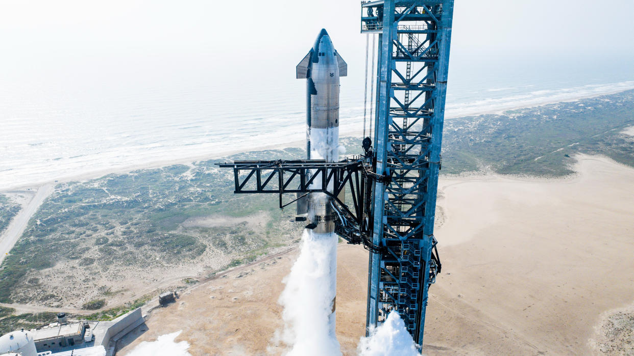  A silver rocket stands next to a huge metallic launch tower, with the ocean in the background. 