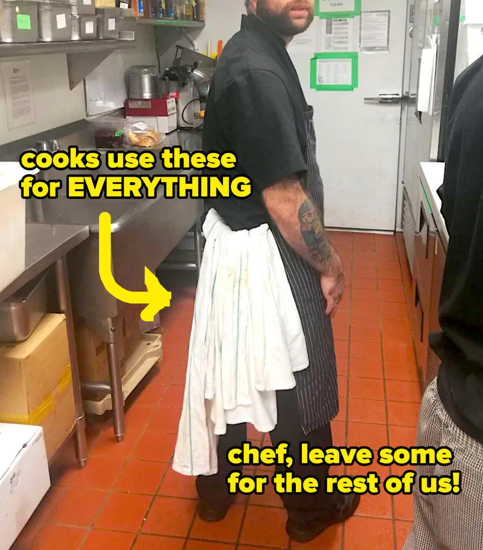 A person is standing in a commercial kitchen, wearing a black shirt, dark-striped apron, and a white towel draped over their back