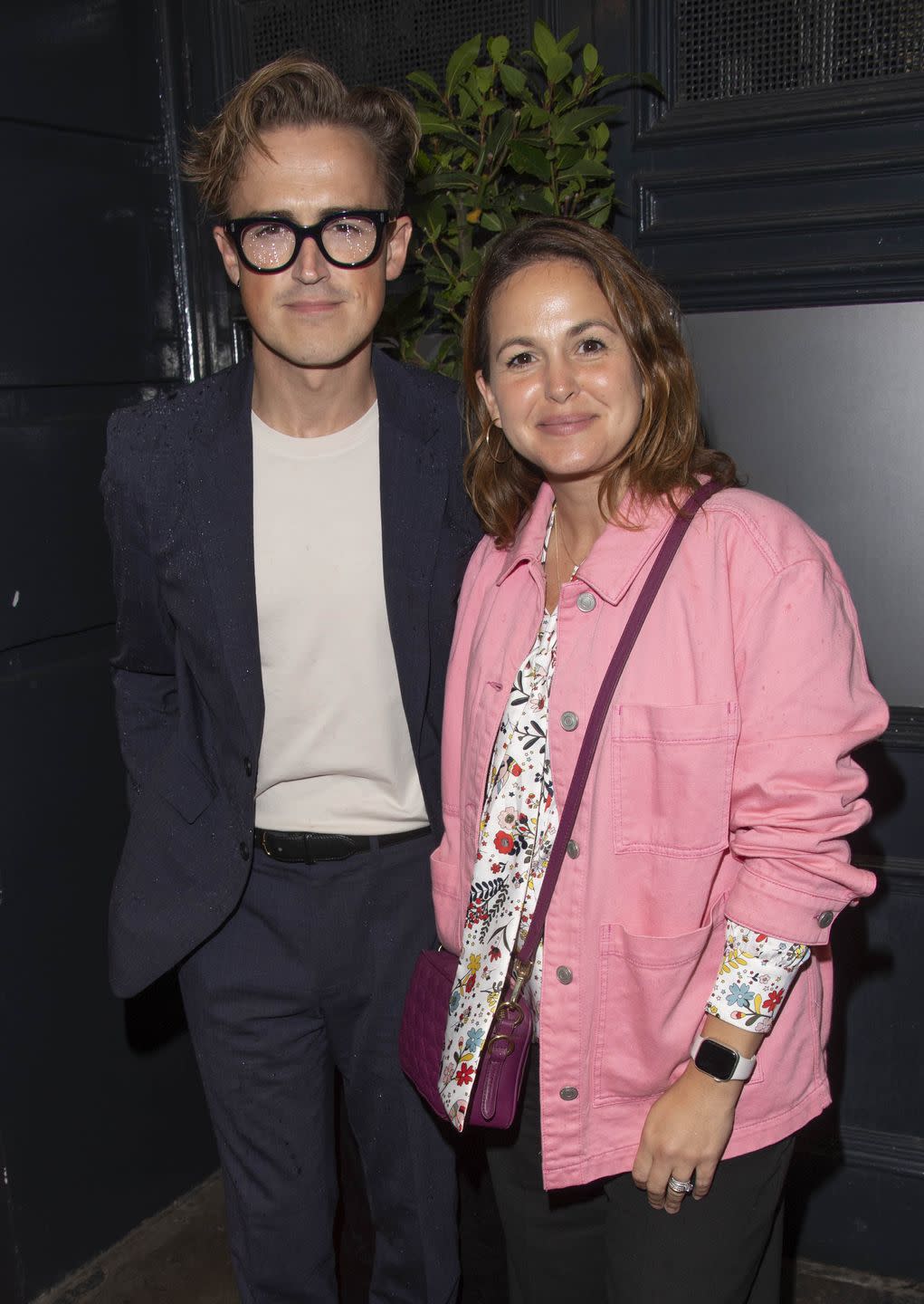 tom fletcher, a man wearing a white shirt and navy suit with thick black glasses, and giovanna fletcher, a woman with short brown hair wearing a pink demin jacket, pose and smile together in front of a leafy plant
