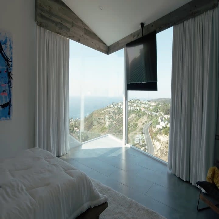 The bedroom has a TV hanging down from the ceiling and the room has huge ceiling to floor windows with an ocean view