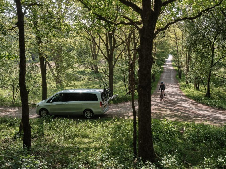 A van driving down a pathway surrounded by trees. There's a biekr nearby.