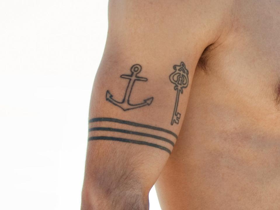 surfer dude with a an anchor key and three band tattoo looking out at the ocean from the beach