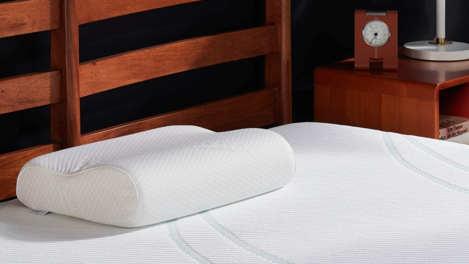 Tempur-pedic neck pillow on a mattress. There is no bedding. The bed, nightstand and clock nearby are all a deep wood.