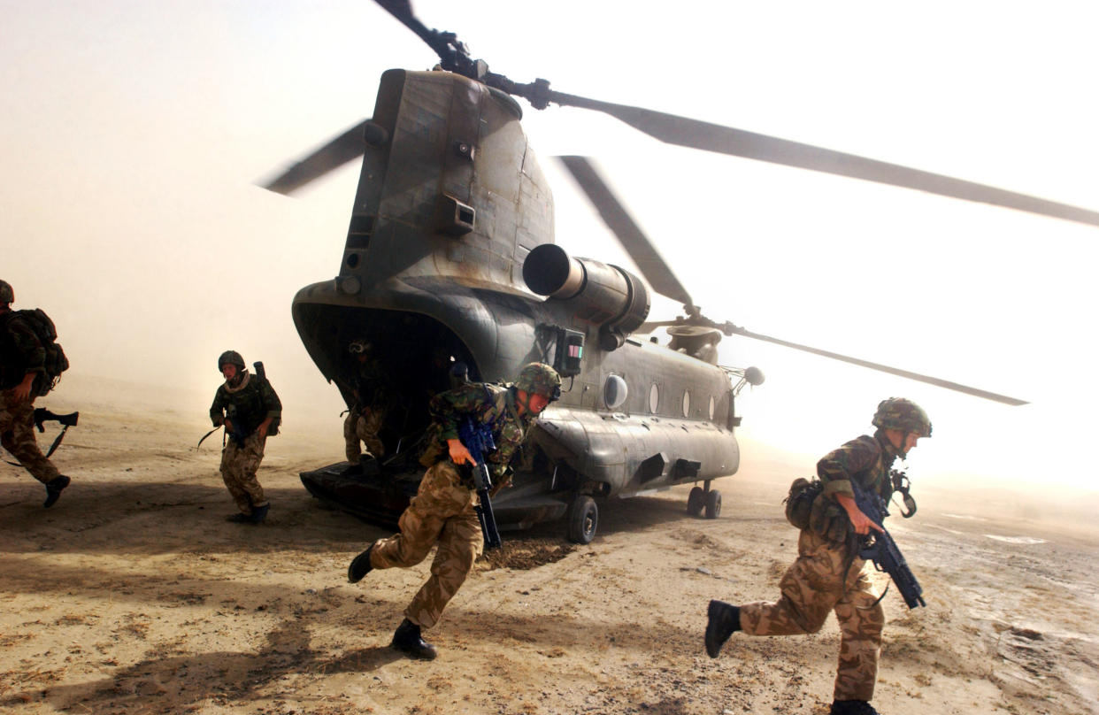 Four military personnel wearing camouflaged clothing and helmets hold guns as they run near the back of a large, two-propellor helicopter resting on sandy ground.