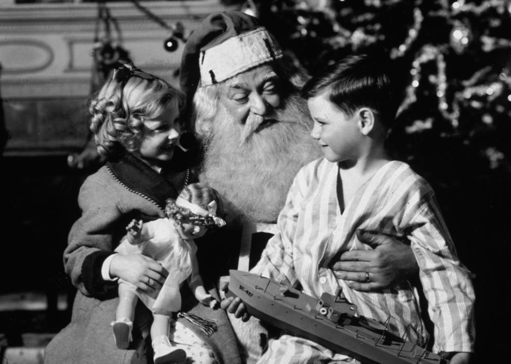 A small boy and girl sit with Santa Claus in front of a Christmas tree.