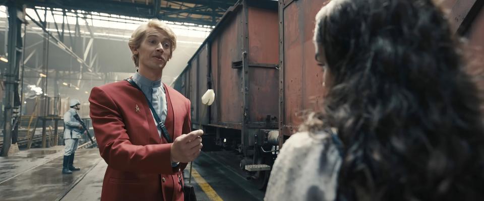 Tom Blyth, in a formal red outfit, holding a flower while interacting with a woman inside an industrial setting in a scene from the film "The Ballad of Songbirds & Snakes"