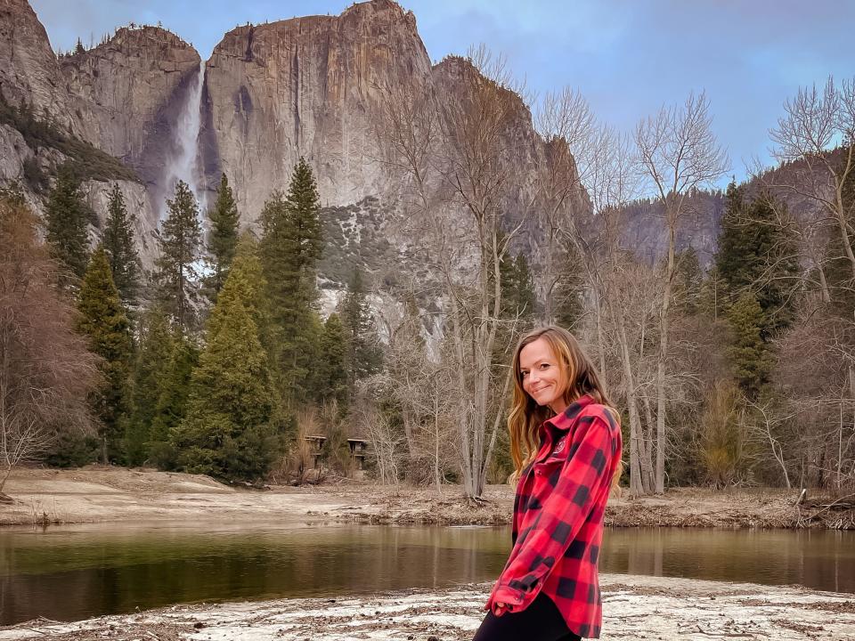 Emily, wearing a red flannel, black leggings, socks, and boots, poses near a small body of water and rock formations at Yosemite National Park.