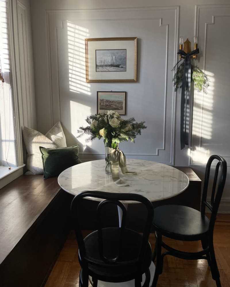 Art is hung on the walls with decorative moldings, next to a marble-topped tulip table and wooden chairs, and a wooden window bench in the room.