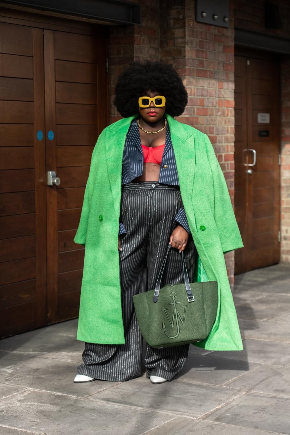 Stephanie Yeboah wears loose pinstripe pants with a cropped pinstripe shirt and red bandeau under a green trench coat.