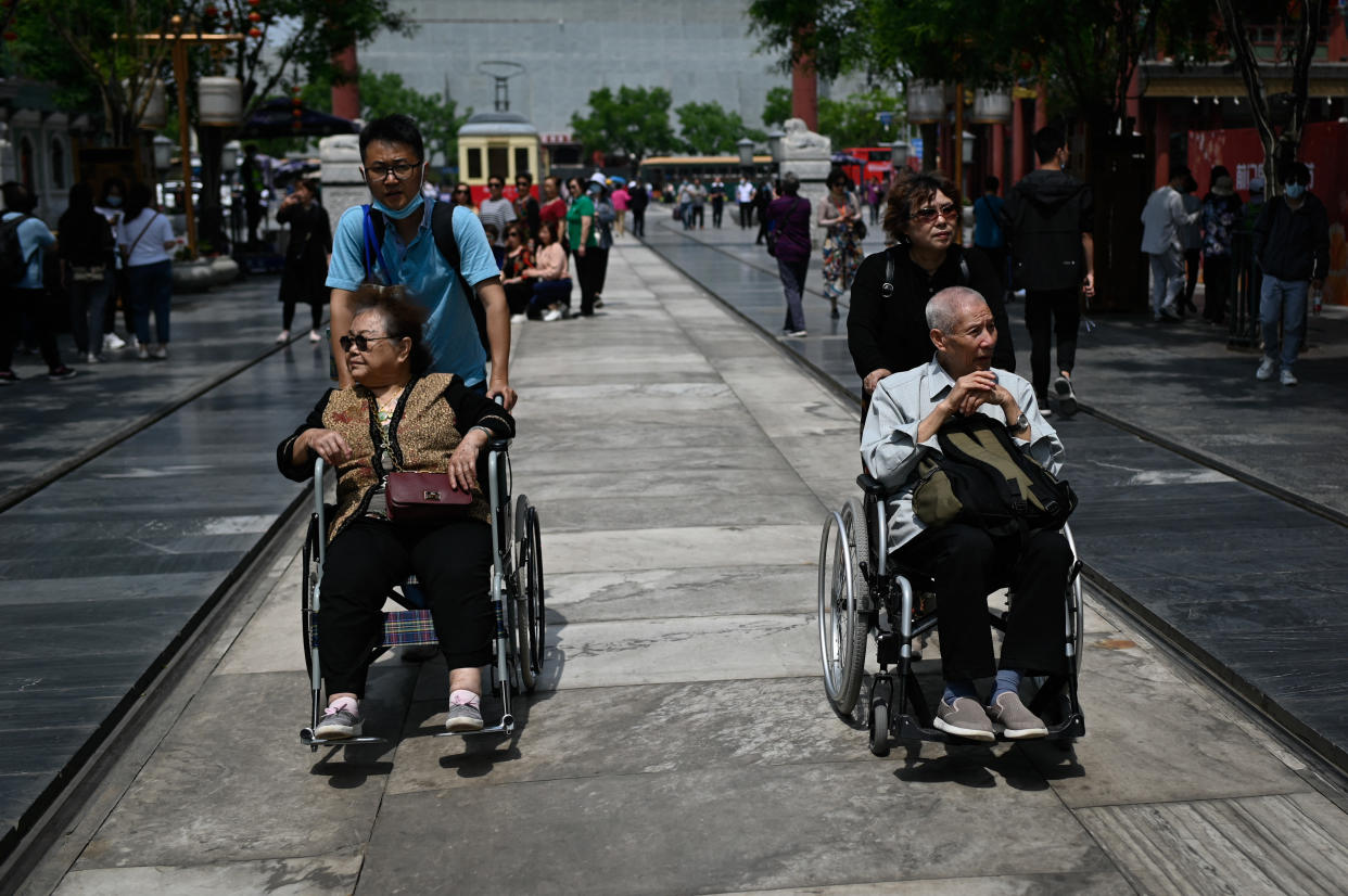 An elderly man and woman are pushed in wheelchairs beside tram tracks on a street in Beijing.