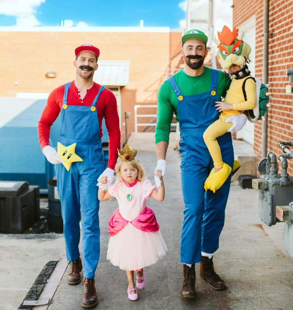 A man dressed as Mario holds hands with a little girl dressed as Princess Peach. She holds hands with a man dressed as Luigi on her other side, who holds a child dressed as Bowser.