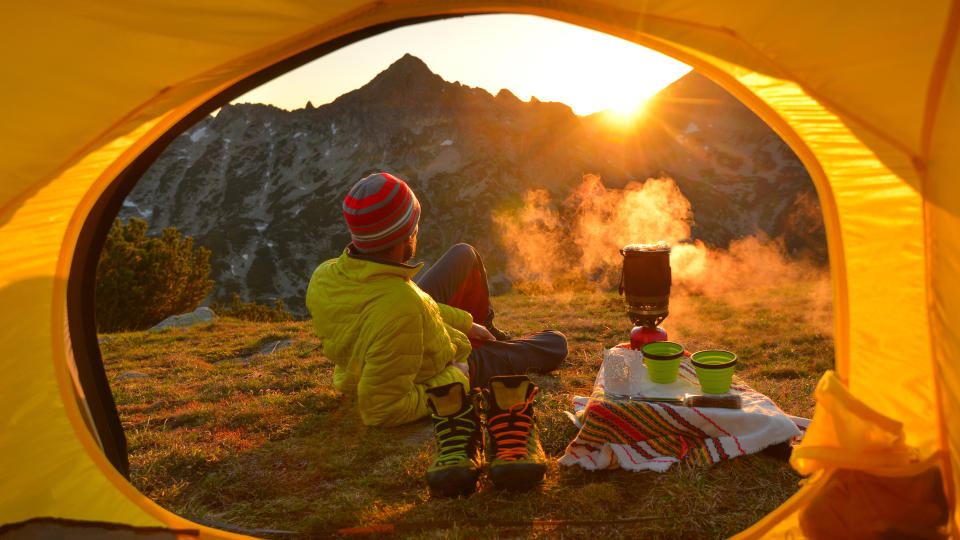 how to get your camping gear ready for summer: a man cooking on a stove outside his tent at sunset