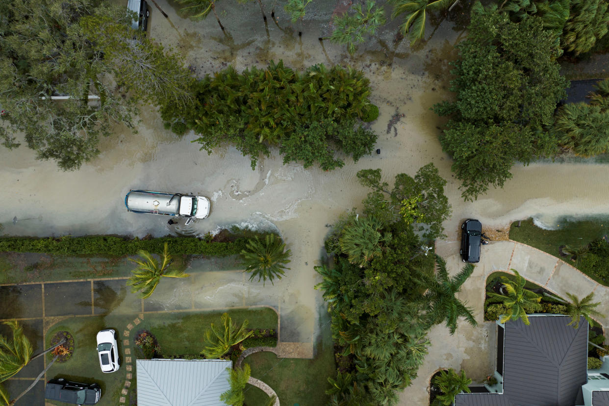 A truck marooned in muddy water along a street seen from the air, with floodwater creeping into adjacent parking lots.