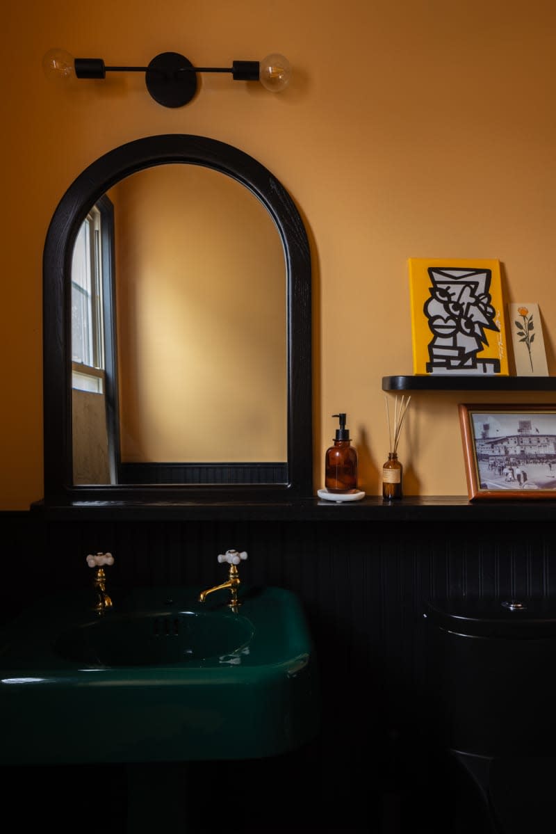 Half-round mirror above green pedestal sink with dual faucets in gold bathroom.