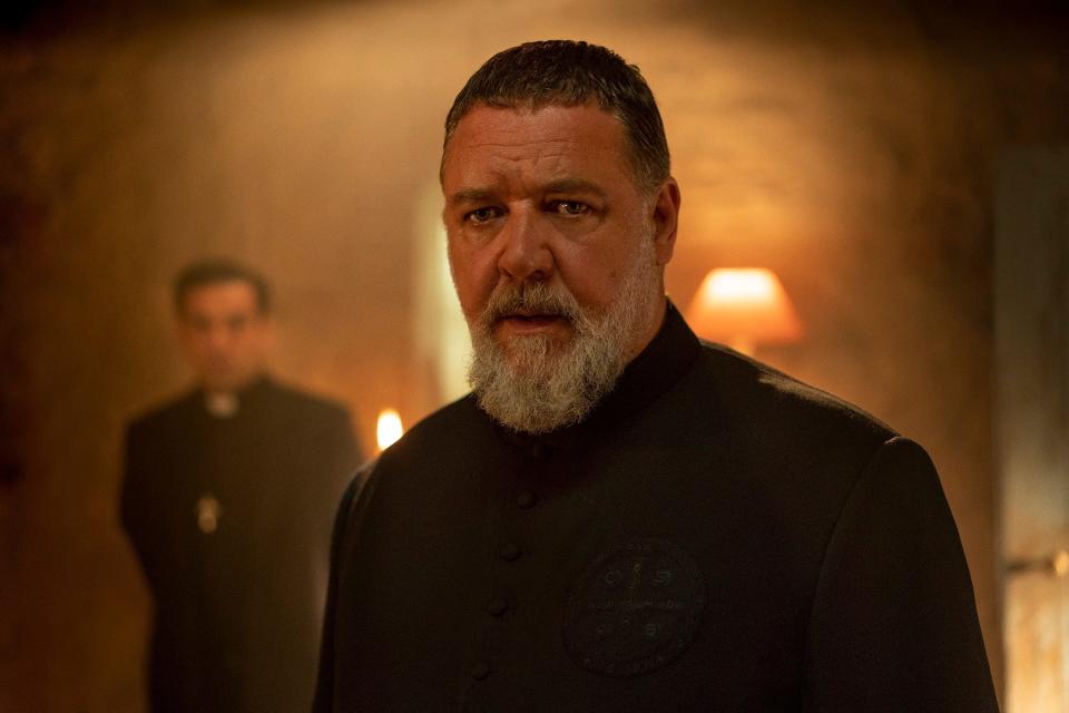 Russell Crowe as Father Gabriele Amorth stands in the foreground while another priest and a lamp are in the background.