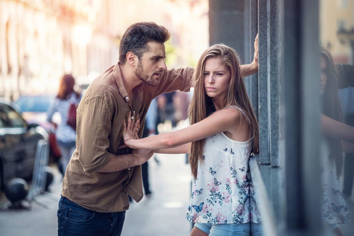 Angry man talking towards woman as she is pushing him away, having a disagreement, she is leaning on the outside of a building, she is looking away from him with a grimace, sidewalk and street with buildings on other side of street, blurred in the backgro