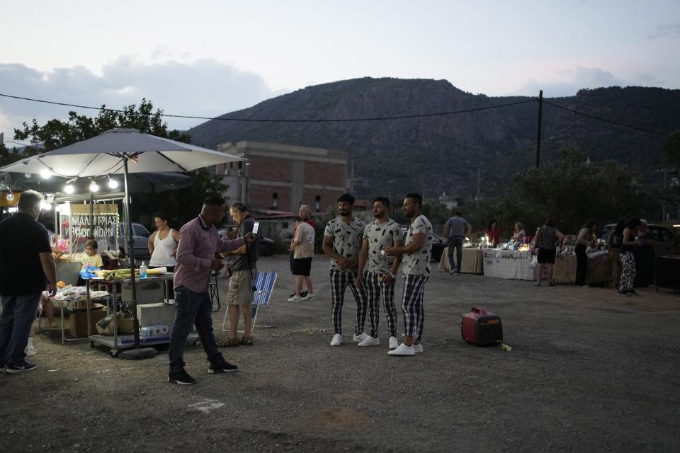 Three men pose for a photograph at a flea market during a festivity, known as Panigiri, in Hasia, northwestern Athens, Greece, Sunday, Aug. 14, 2022. The Dormition of the Virgin Mary (or Mother of God as the Greeks usually refer to her) is celebrated on Aug. 15. The religious event is coupled with midsummer festivities, known as Panigiria, that often last more than a day with music, culinary feasts and, in many cases, flea markets. (AP Photo/Thanassis Stavrakis)
