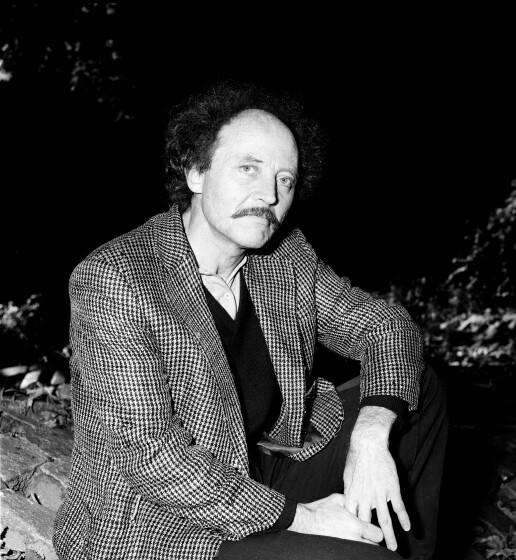 Sylvere Lotringer, in a sweater and checked blazer, is seen sitting with his hands on his knee in a black and white photo