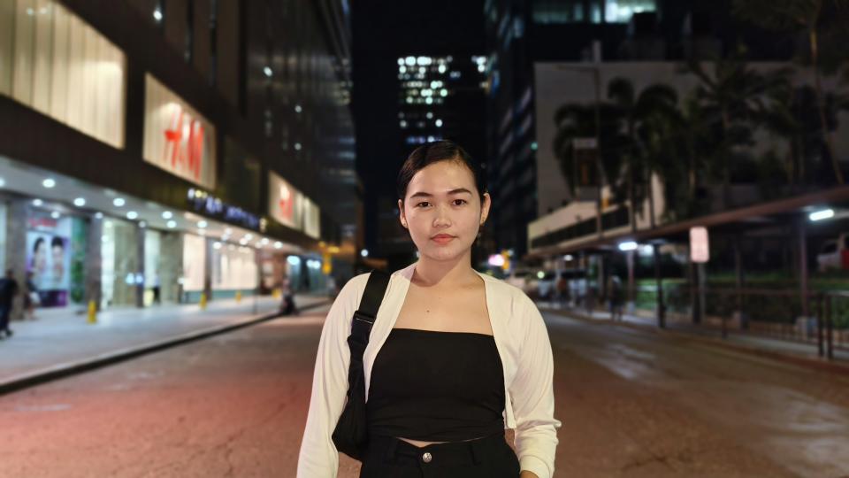 A woman standing alone on a city street at night, surrounded by tall buildings and illuminated by streetlights