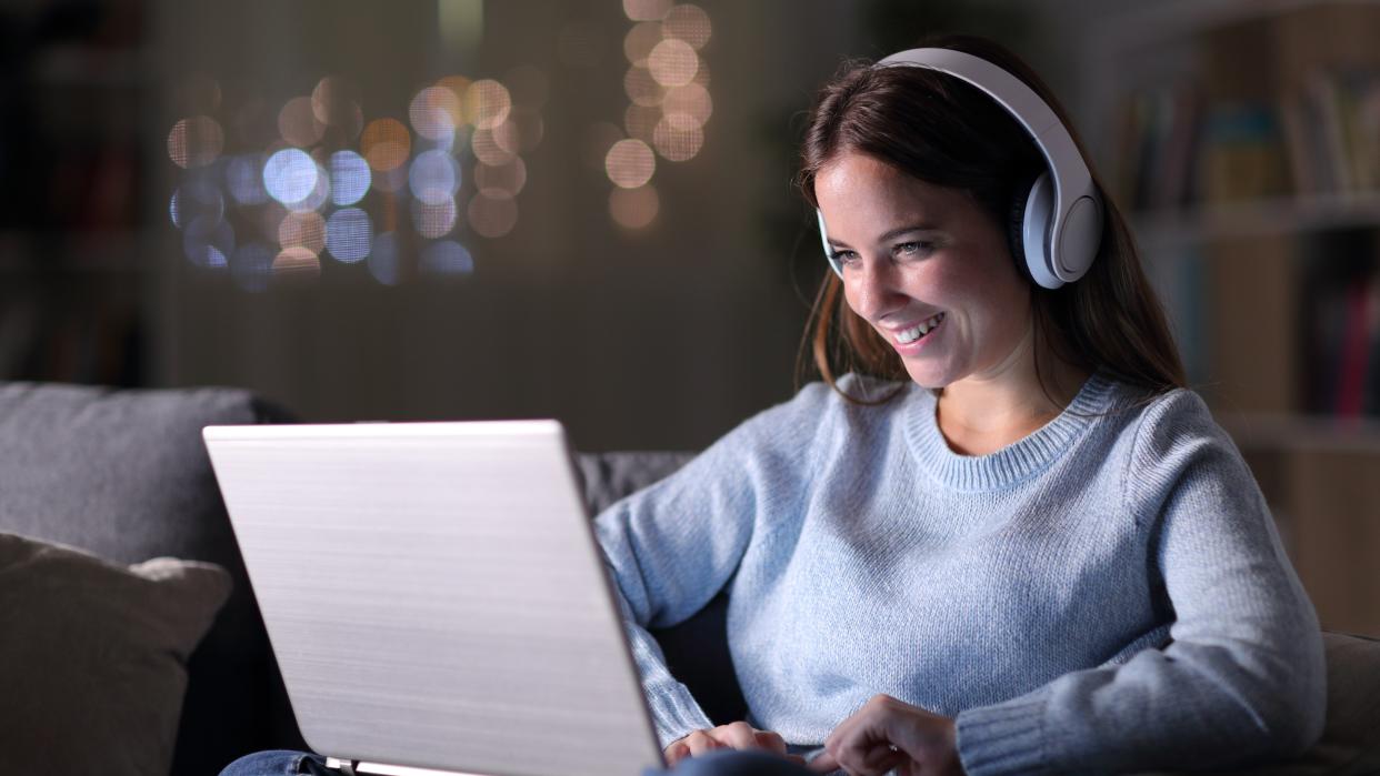  A woman sitting on a coach and on her laptop, wearing headphones, at night time. 