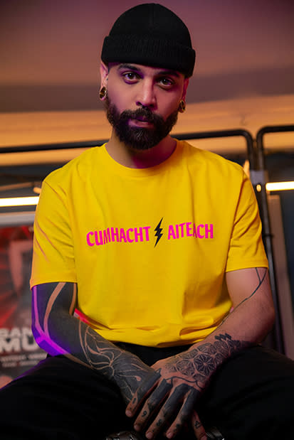 A model wearing queer GCN merch - a yellow t-shirt reading "Cumhacht Aiteach"