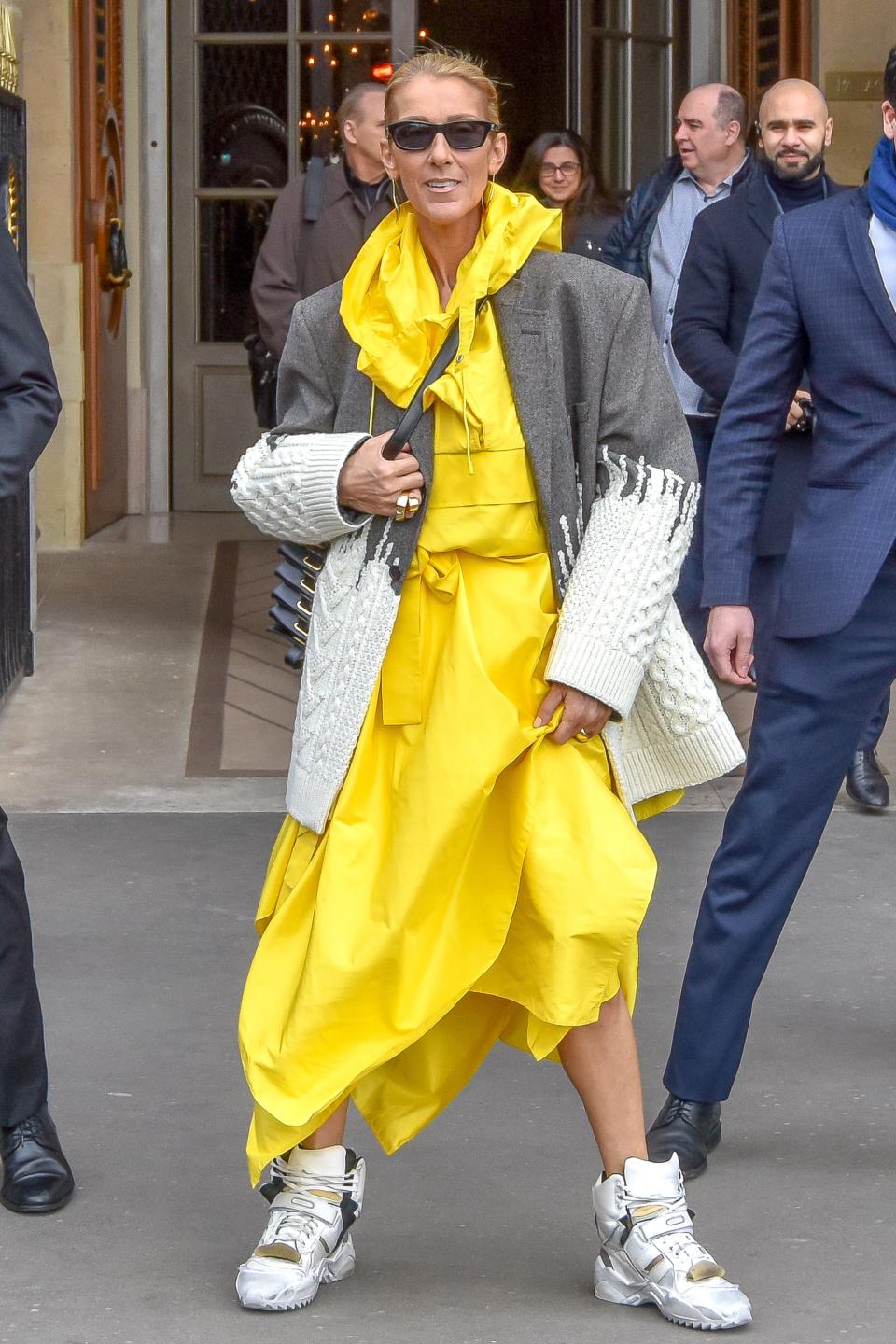 Celine in a yellow a-symmetrical raincoat dress under a grey and white knitted blazer. She's wearing sunglasses and white chunky sneakers.