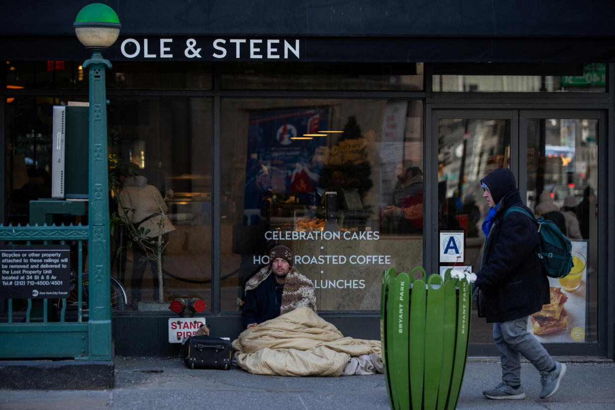At an Ole and Steen coffee shop by the subway, under a sign saying Celebration Cakes and Hot Roasted Coffee, a man in a wool hat and a hard stare sits with a small suitcase by his side, under a heap of bedding.