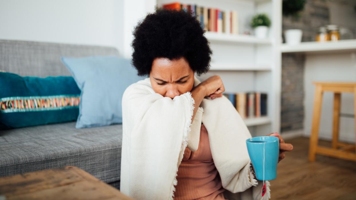  Woman can be seen sat on a wooden floor coughing into her elbow while she holds a cup of tea in a blue mug in the other hand. The background is blurred, however, she is sat next to a wooden coffee table and a grey couch with blue cushions. There is also a bookshelf behind her and a wooden stool against a kitchen counter to the right. 