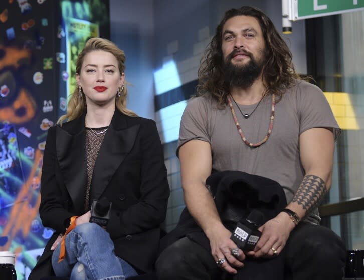 A blond woman and a large bearded man hold microphones while sitting and waiting to answer questions