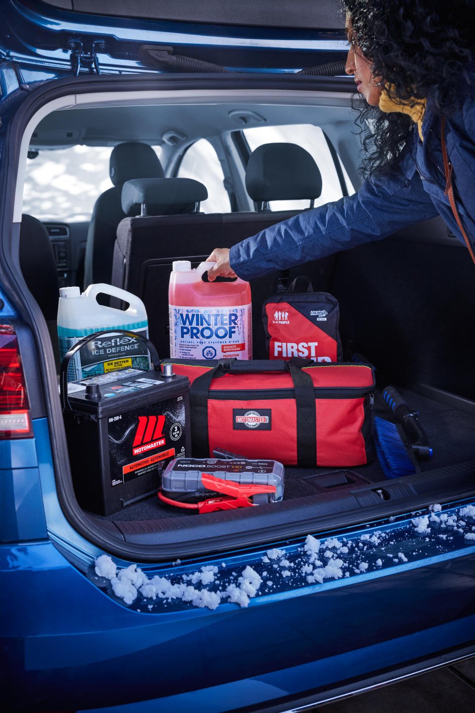 Woman putting winter driving safety essentials from Canadian Tire in her trunk, including winter washer fluid, a booster pack, winter battery, first aid kit and winter safety kit for roadside emergencies