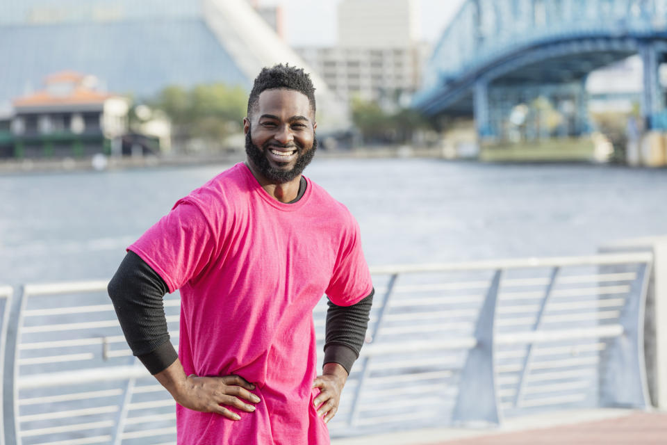 A person poses confidently with hands on hips by a river, wearing a casual layered look with a short-sleeve shirt over long sleeves