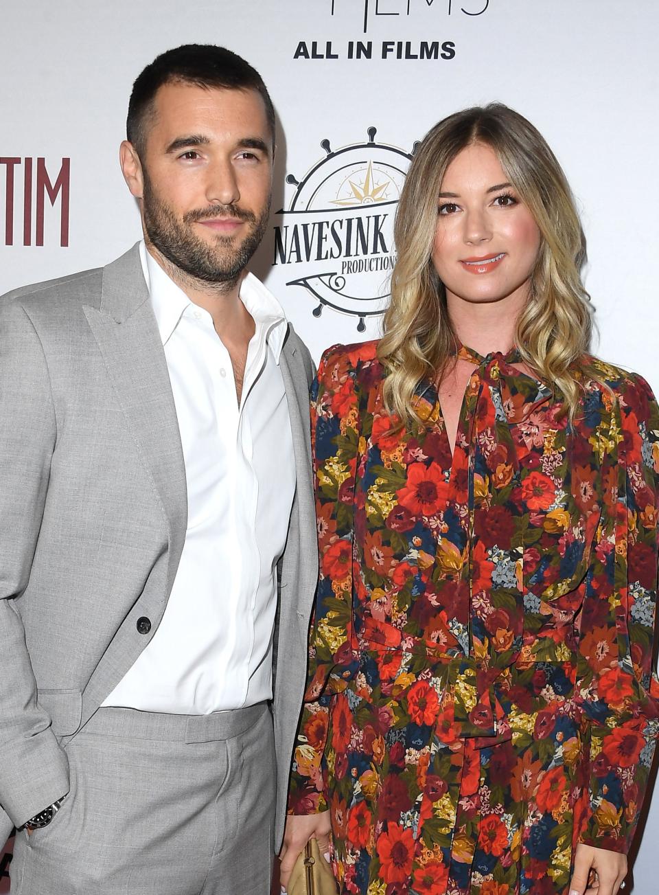 Man in a gray suit and woman in a floral dress standing together at an event with a logo backdrop